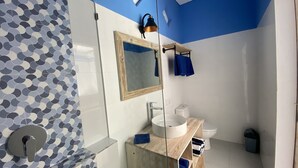 A modern bathroom with a blue and white tiled wall, a wooden vanity, a white sink, a mirror, and a shower area. - Kura Kura Divers Lodge (Karangasem)