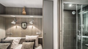 Een moderne slaapkamer met een houten hoofdbord, een bed met witte lakens en meerdere kussens, twee wandlampen en een douchecabine van glas.