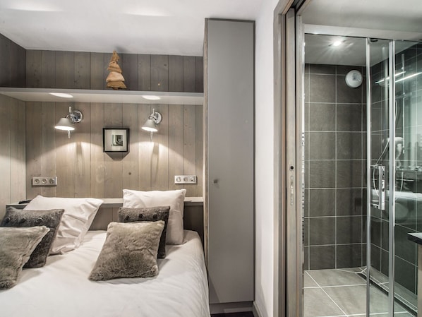 A modern bedroom with a wooden headboard, a bed with white linens and multiple pillows, two wall-mounted lights, and a glass shower enclosure.