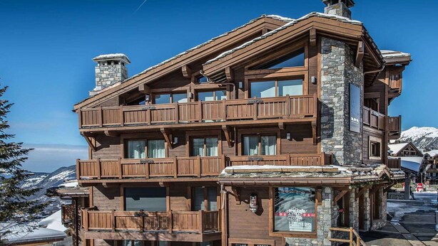 Een meerhoog gebouw van hout met balkons, stenen schoorstenen en uitzicht op besneeuwde bergen.