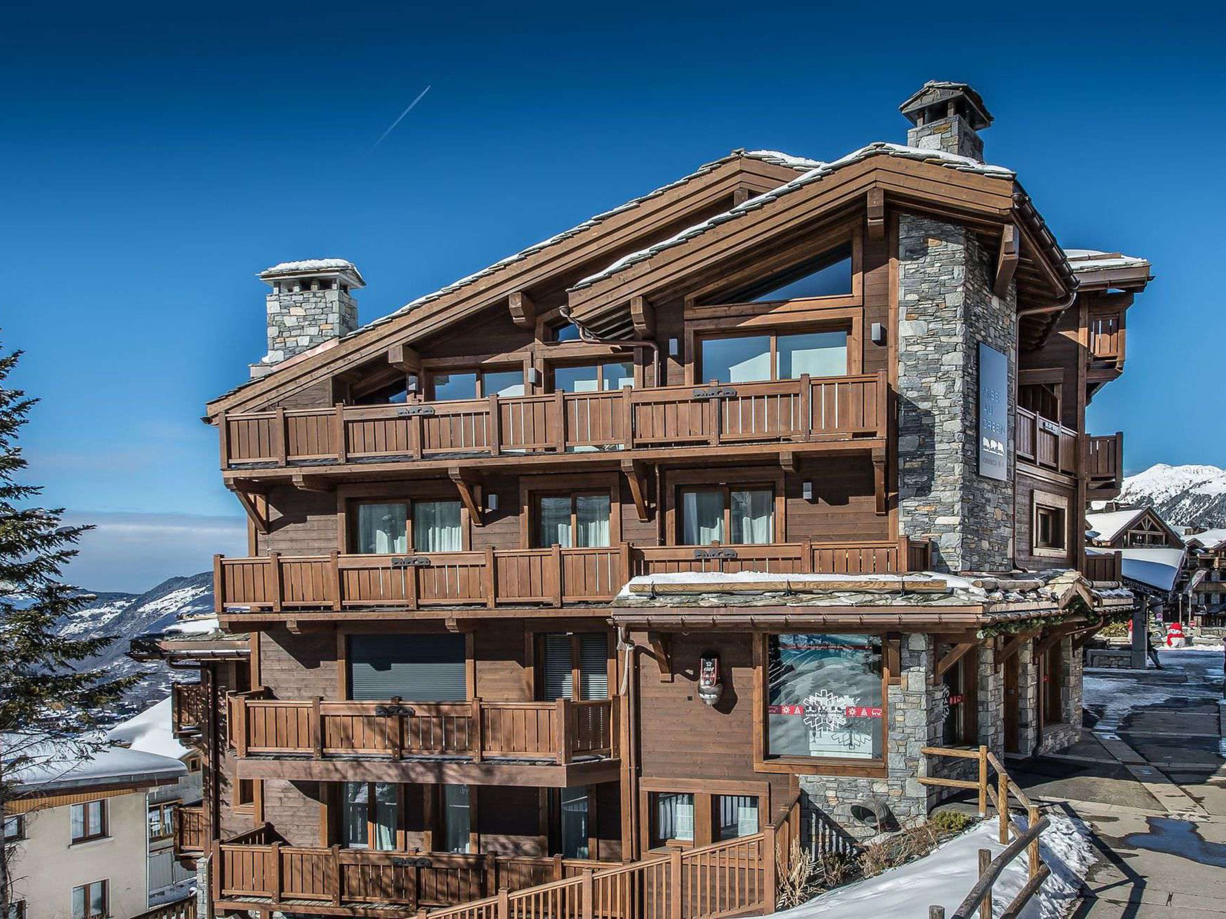 A multi-story wooden building with balconies, stone chimneys, and a view of snow-covered mountains.