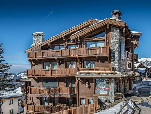 A multi-story wooden building with balconies, stone chimneys, and a view of snow-covered mountains.