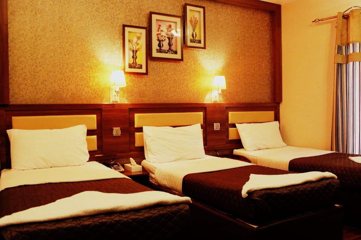 A hotel room with three beds, each with white linens and brown headboards. There are wall-mounted lamps on either side of the beds, and three framed pictures above the beds.