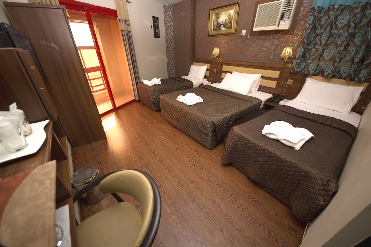 A hotel room with two beds, a wooden floor, a chair, a desk with a lamp, and a painting on the wall.