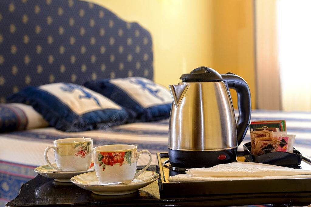 A neatly arranged breakfast setup with a kettle, two cups, and a tray on a bed with patterned pillows.