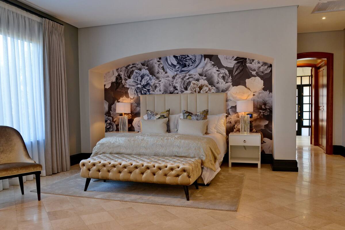 A bedroom with a large bed, a tufted ottoman, a chair, a nightstand with a lamp, and a floral wall mural.