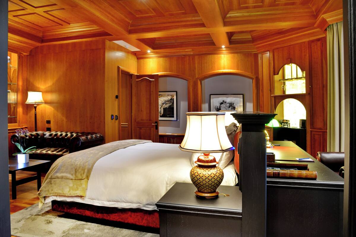 A hotel room with a large bed, a wooden ceiling, a desk with a lamp, and a leather sofa.
