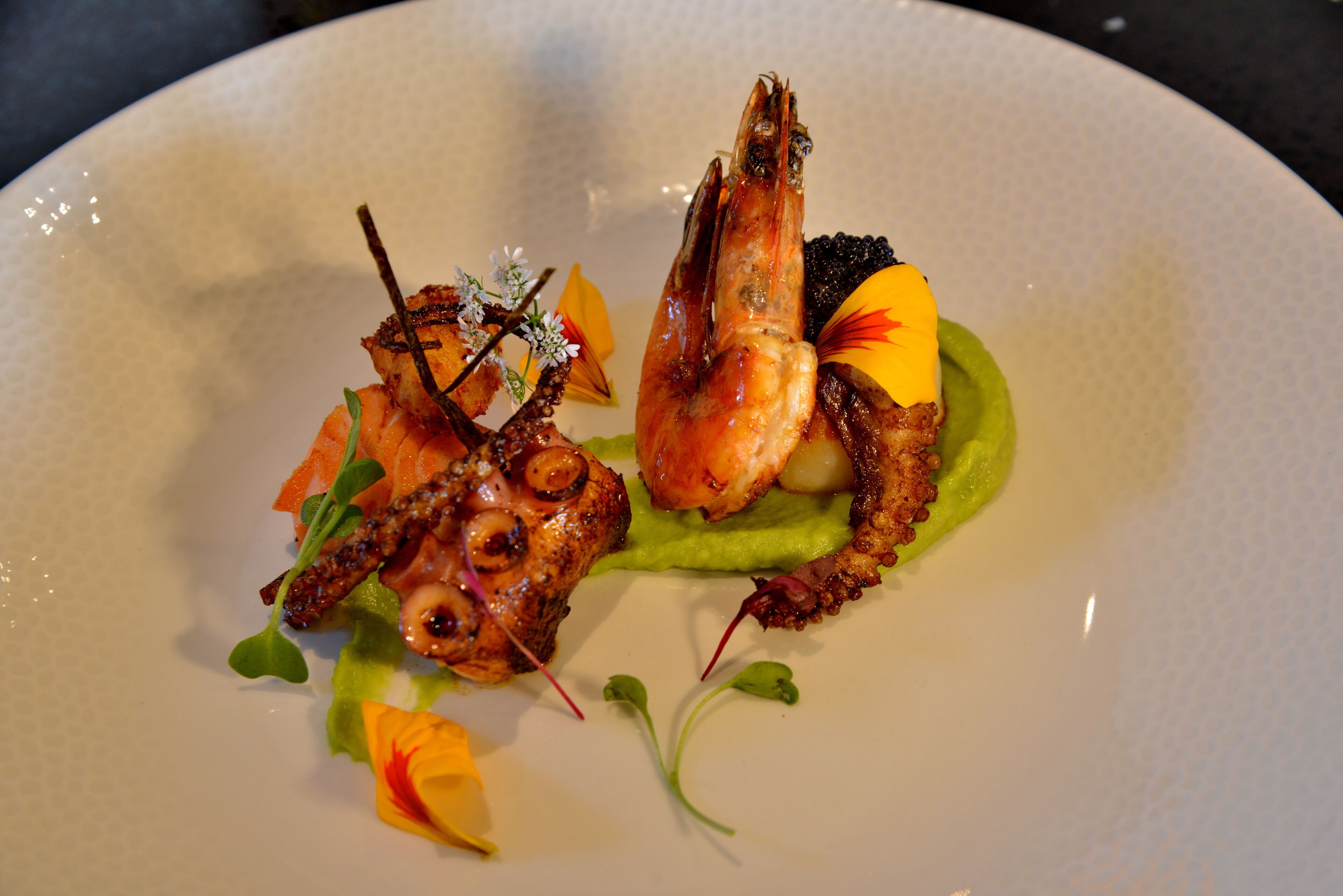 Une assiette de fruits de mer, comprenant peut-être des crevettes et des poulpes, agrémentée de fleurs et d’herbes comestibles.