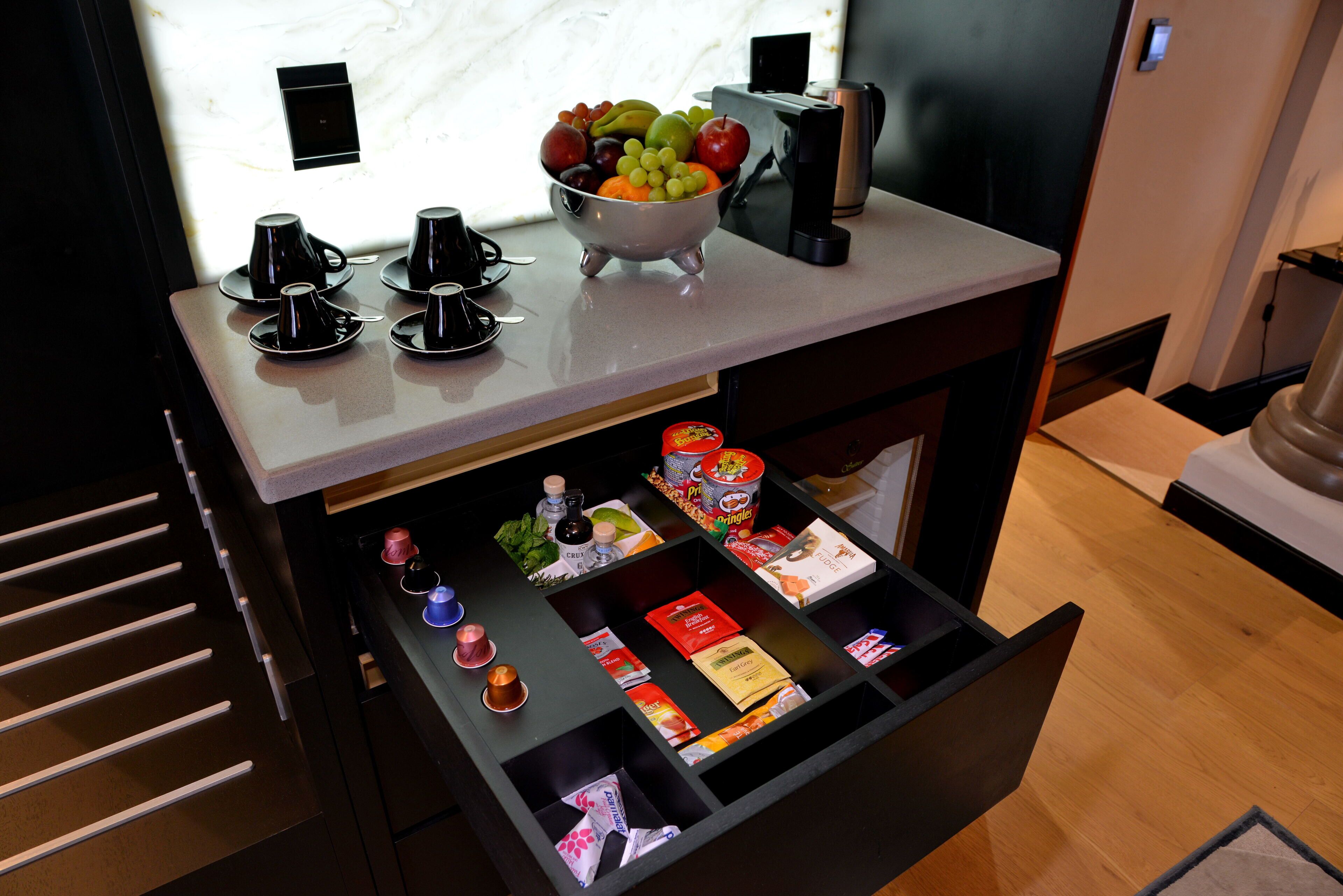 Un mini-bar d’hôtel avec un panier de fruits, une machine à café et divers en-cas.