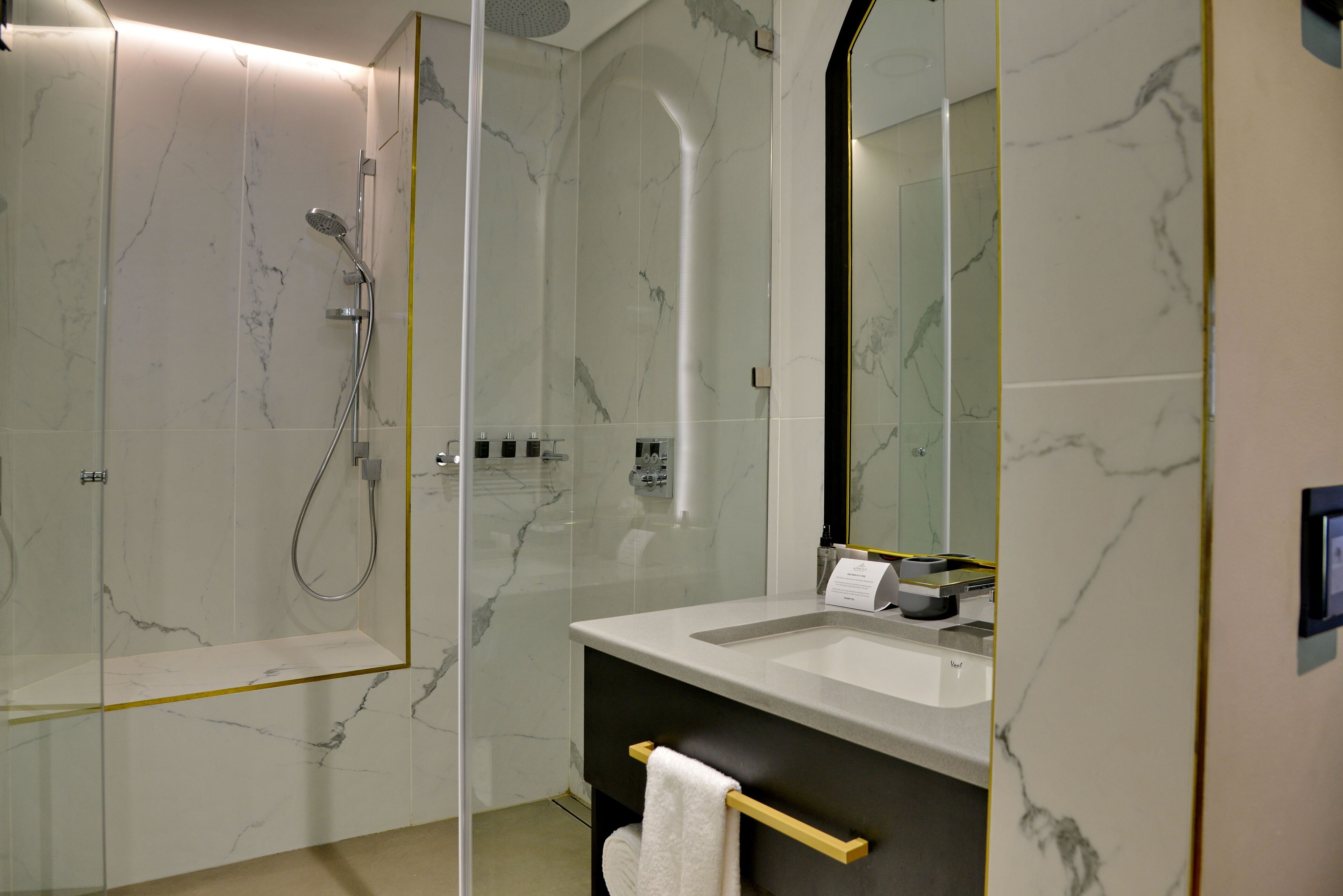 Un baño moderno con ducha envuelta en vidrio, pared con azulejos de mármol, lavamanos con espejo y barra para toallas.