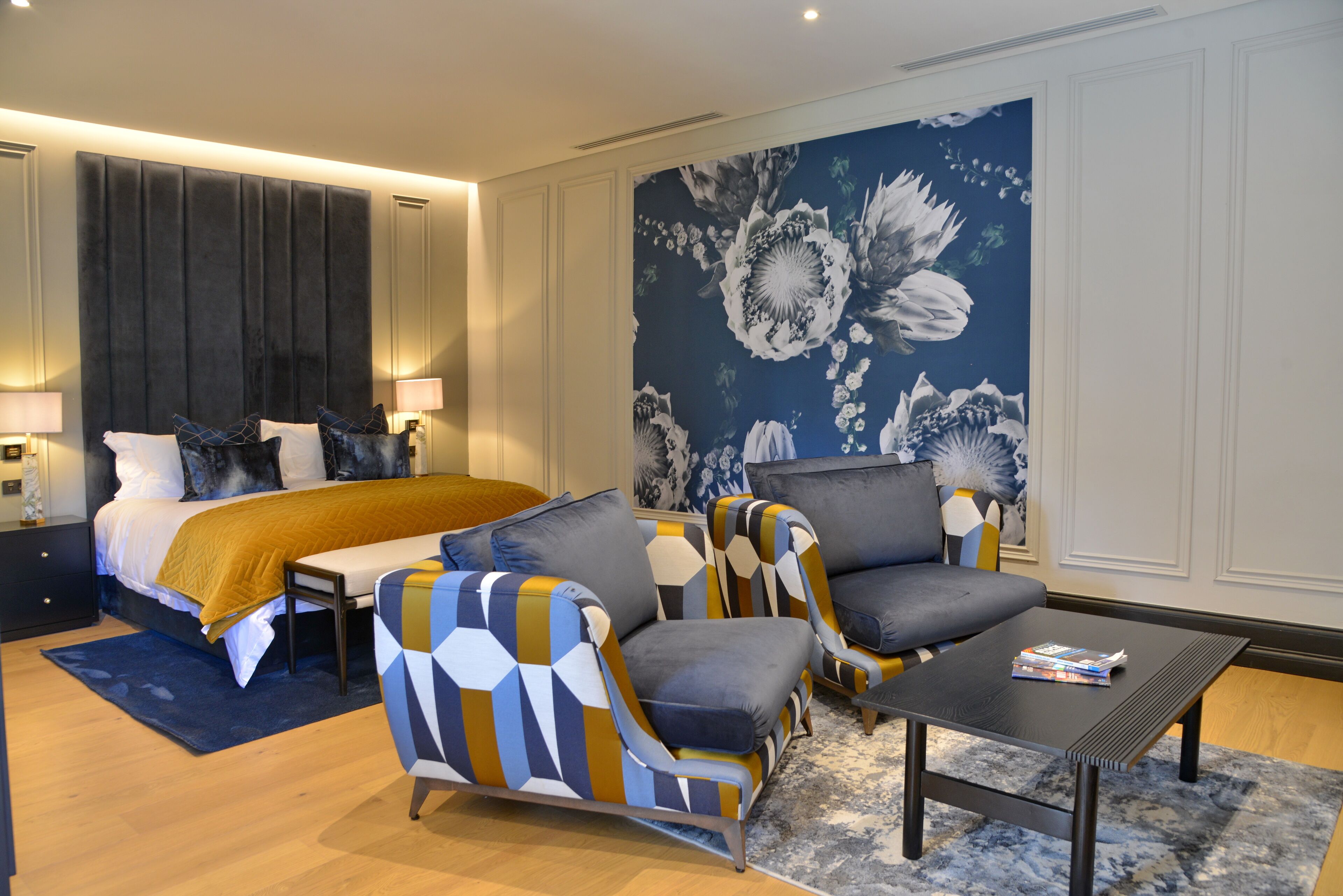 Una habitación de hotel moderna con una cama grande, dos sillones, una mesita de centro y un mural floral en la pared.