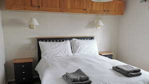 Un dormitorio con cabecera de madera, dos mesitas de noche con lámparas, una cama con ropa blanca y un aparador de madera sobre la cama.