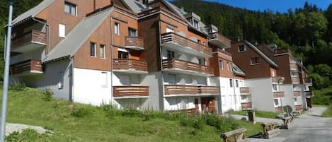 A row of modern multi-story buildings with balconies, set against a backdrop of green hills and a clear sky.
