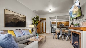 Ein Wohnzimmer mit Sofa, Couchtisch und Kamin. Eine Essecke mit Tisch und Stühlen ist ebenfalls vorhanden. Im Hintergrund ist eine Küche mit Schränken und Geräten zu sehen.