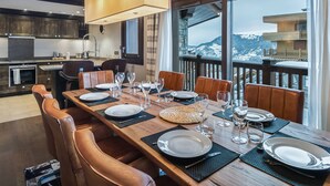 Un coin repas avec une table en bois dressée pour six personnes, une cuisine en arrière-plan et une vue sur les montagnes par de grandes fenêtres.