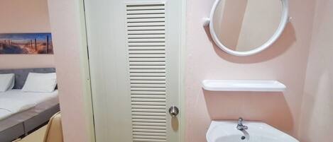 A room with a door, a bed, a bedside table, a round mirror, and a sink.