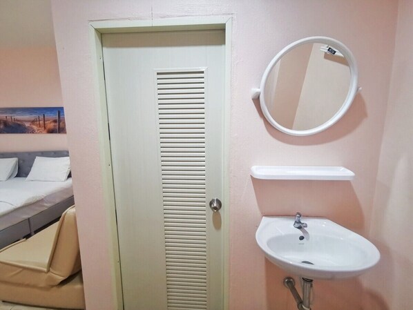A room with a door, a bed, a bedside table, a round mirror, and a sink. - Pratubjai Hotel (Sattahip)