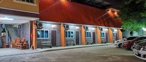 Un edificio con varias habitaciones, cada una con ventana y unidad de aire acondicionado. Hay sillas y mesas afuera, y un coche estacionado cerca.