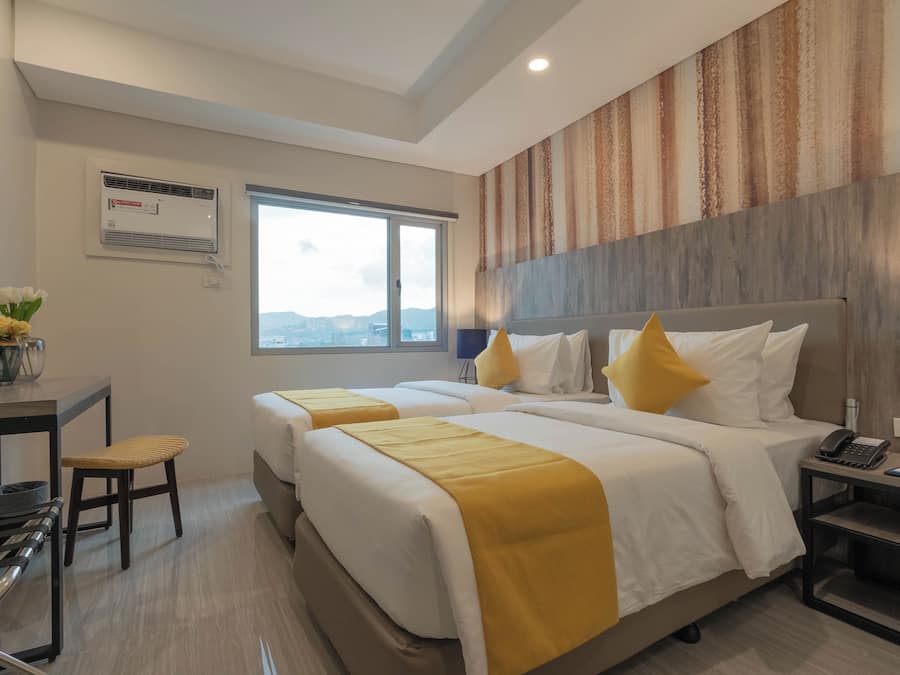 Una habitación de hotel con dos camas, un escritorio y una silla, un ventanal con vistas y un equipo de aire acondicionado.