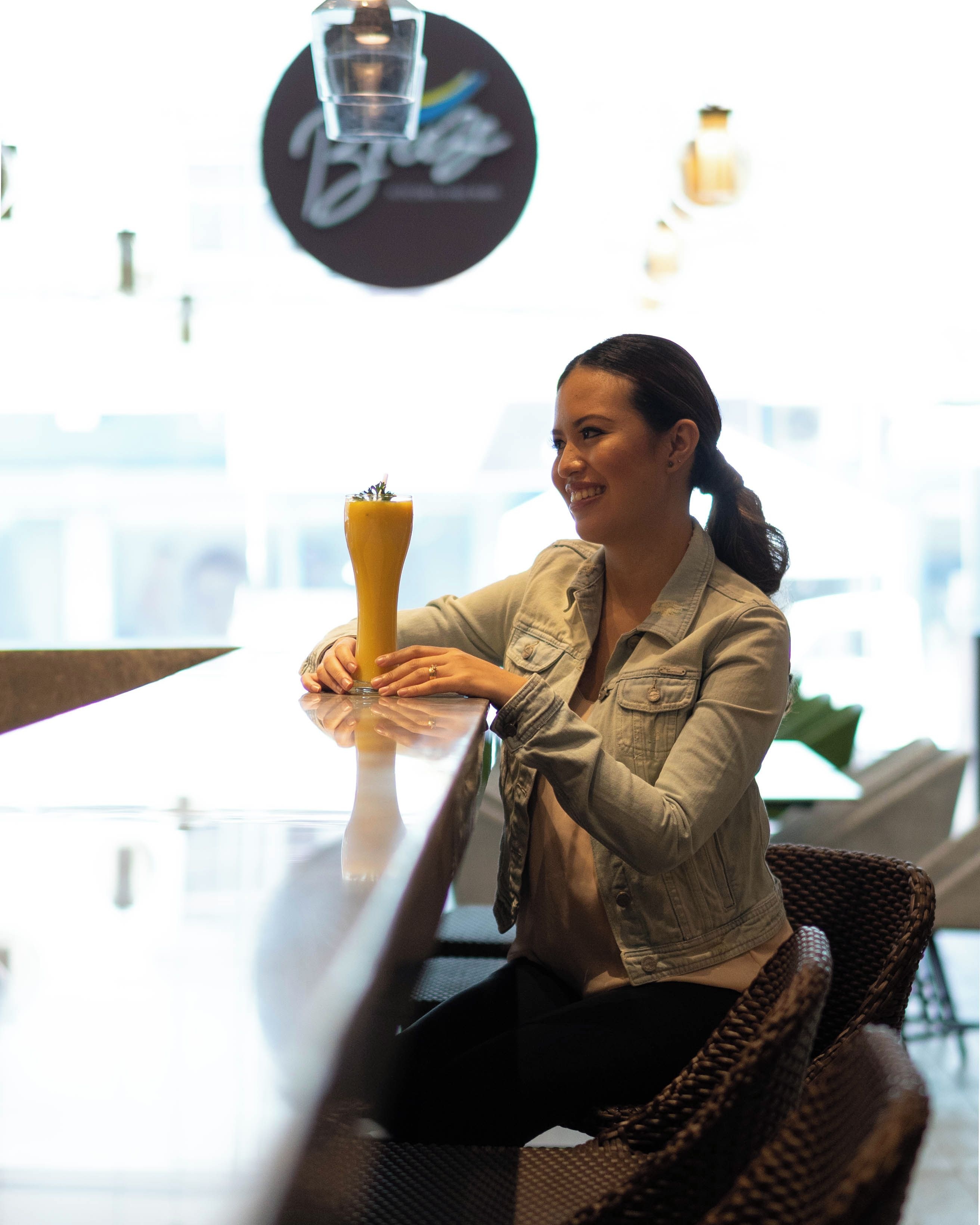 A person seated at a table with a tall glass of a yellow beverage, in a setting with a circular sign above and a window in the background.