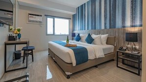 A modern hotel room with a large bed, a desk with a chair, a small table, and a nightstand with a lamp.