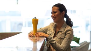 A person sitting at a table with a drink, in a well-lit indoor setting with a visible sign in the background.