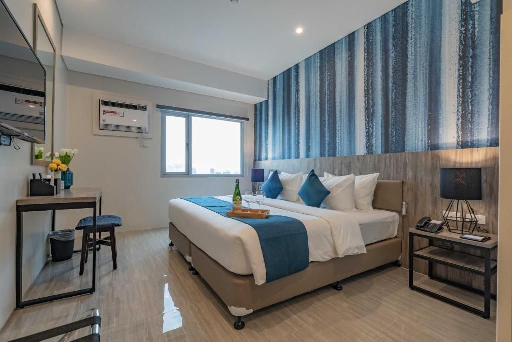 Una habitación de hotel moderna con una cama grande, un escritorio con una silla, una mesita pequeña y una mesita de noche con una lámpara.