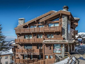 A multi-story wooden building with balconies, stone chimneys, and a view of snow-covered mountains.