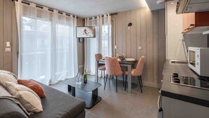 A modern living room with a sofa, a dining table with chairs, a TV mounted on the wall, and a kitchen area with a microwave. - Cozy apartment in Courchevel with spacious bedroom and parking (Courchevel 1650)