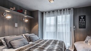 A modern bedroom with a large bed, a wooden headboard, and a wall-mounted shelf with decorative items. - Cozy apartment in Courchevel with spacious bedroom and parking (Courchevel 1650)