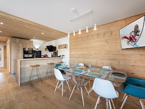Una cocina moderna con detalles de madera, un comedor con mesa y sillas de vidrio, y un cuadro grande en la pared.