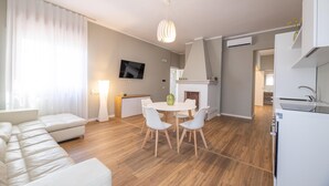 Uma sala de estar moderna com sofá, mesa de jantar com cadeiras, TV na parede e área de cozinha com fogão.