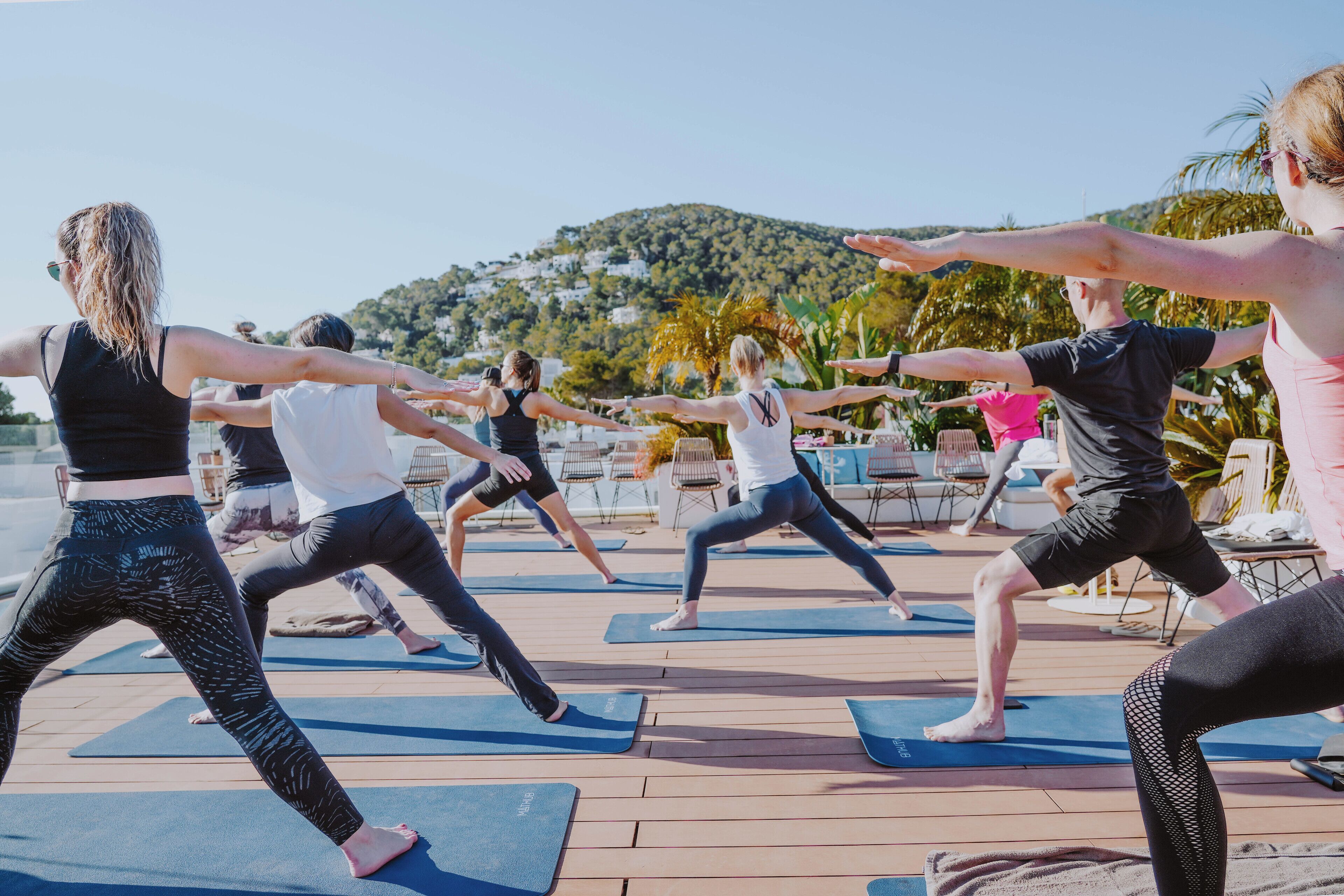 A group of people performing yoga on mats outdoors with a scenic view of hills and palm trees in the background.