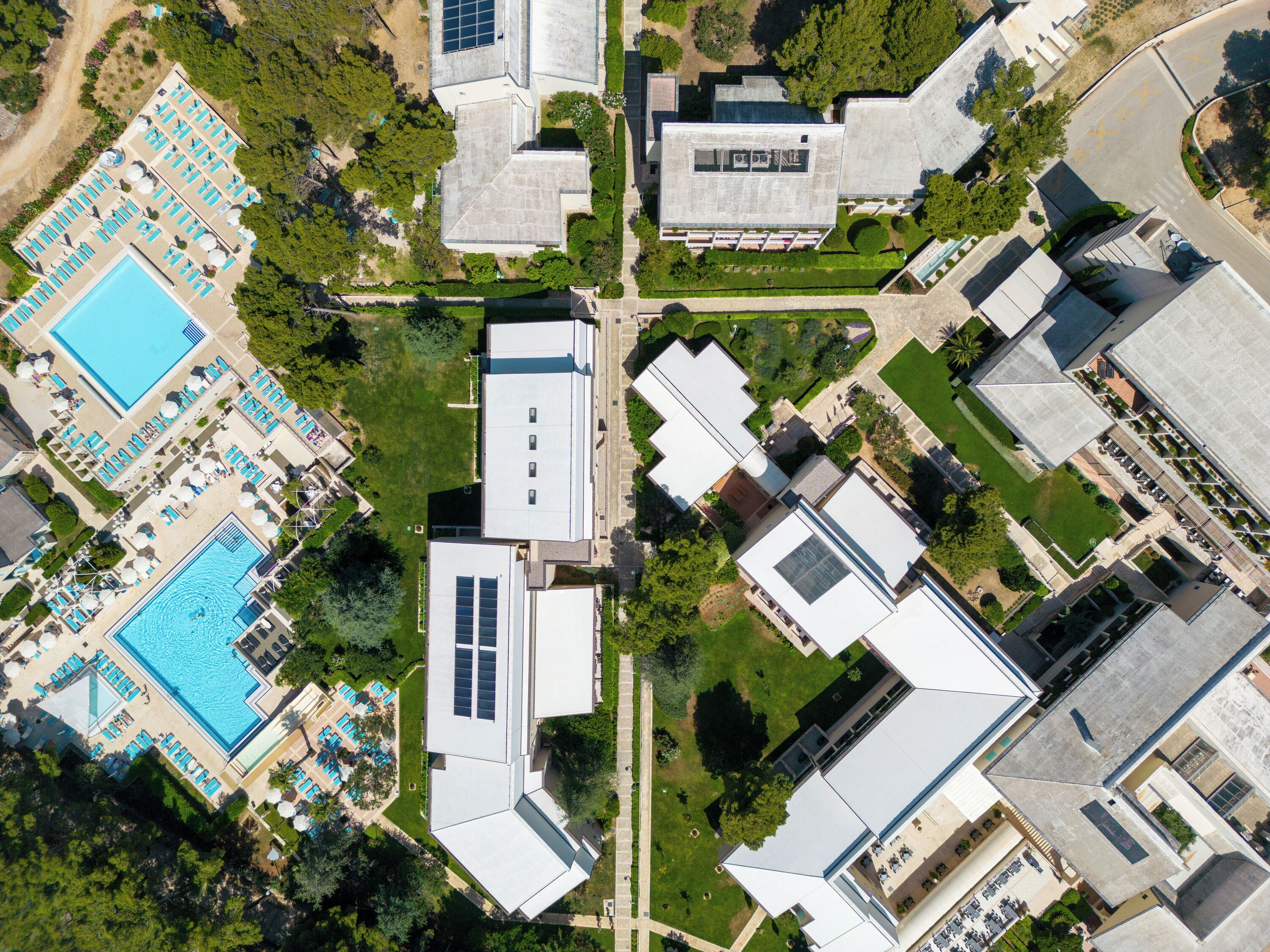Vista aérea de um hotel com vários edifícios, incluindo uma grande área de piscina com espreguiçadeiras e um edifício separado com telhado plano.