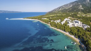Vista aerea di una zona costiera con una spiaggia limpida, uno yacht e un resort con edifici e alberi.