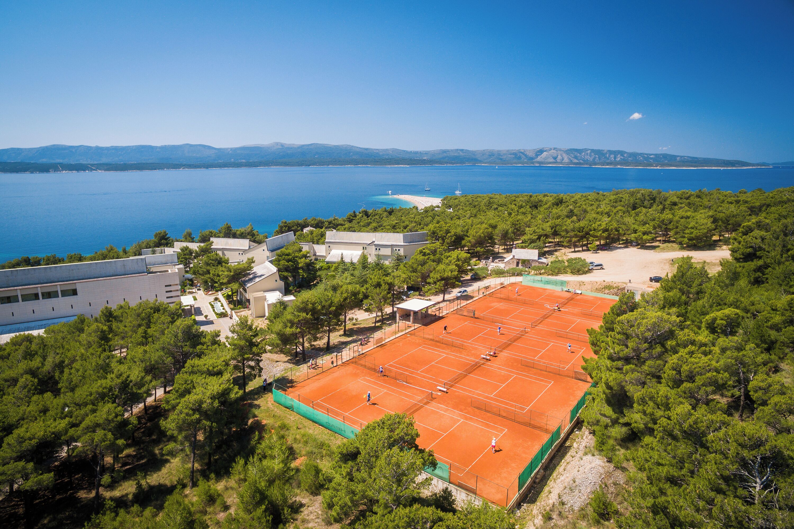 Una cancha de tenis rodeada de árboles, con vistas al mar y montañas al fondo.