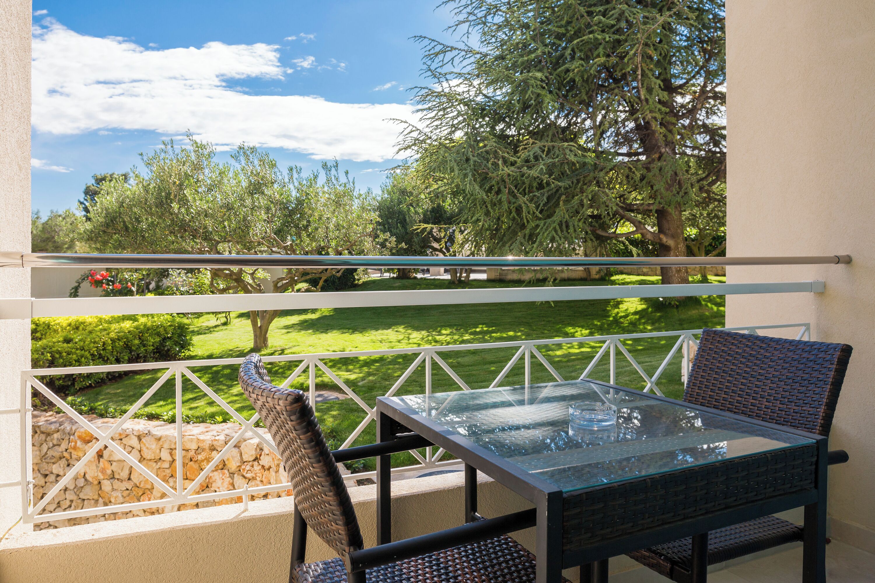 Un balcon avec une table en verre et des chaises en osier, donnant sur un jardin avec des arbres et un mur de pierre.