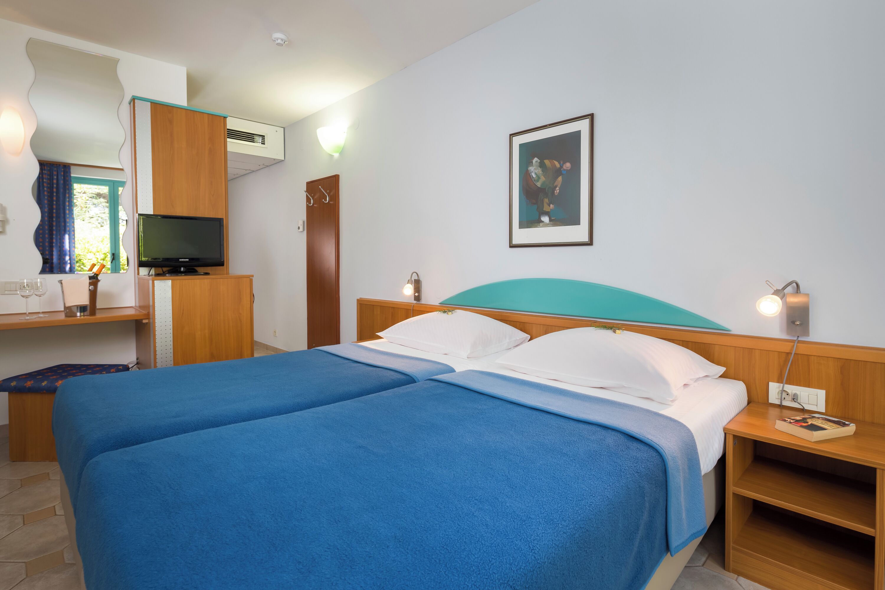A hotel room with two beds, a desk with a TV, a framed picture on the wall, and a window with curtains.