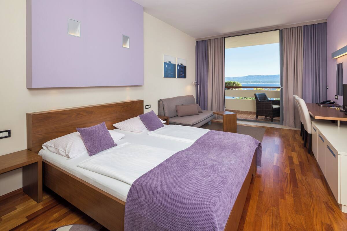 A hotel room with a large bed, a desk, a chair, and a balcony with a view.