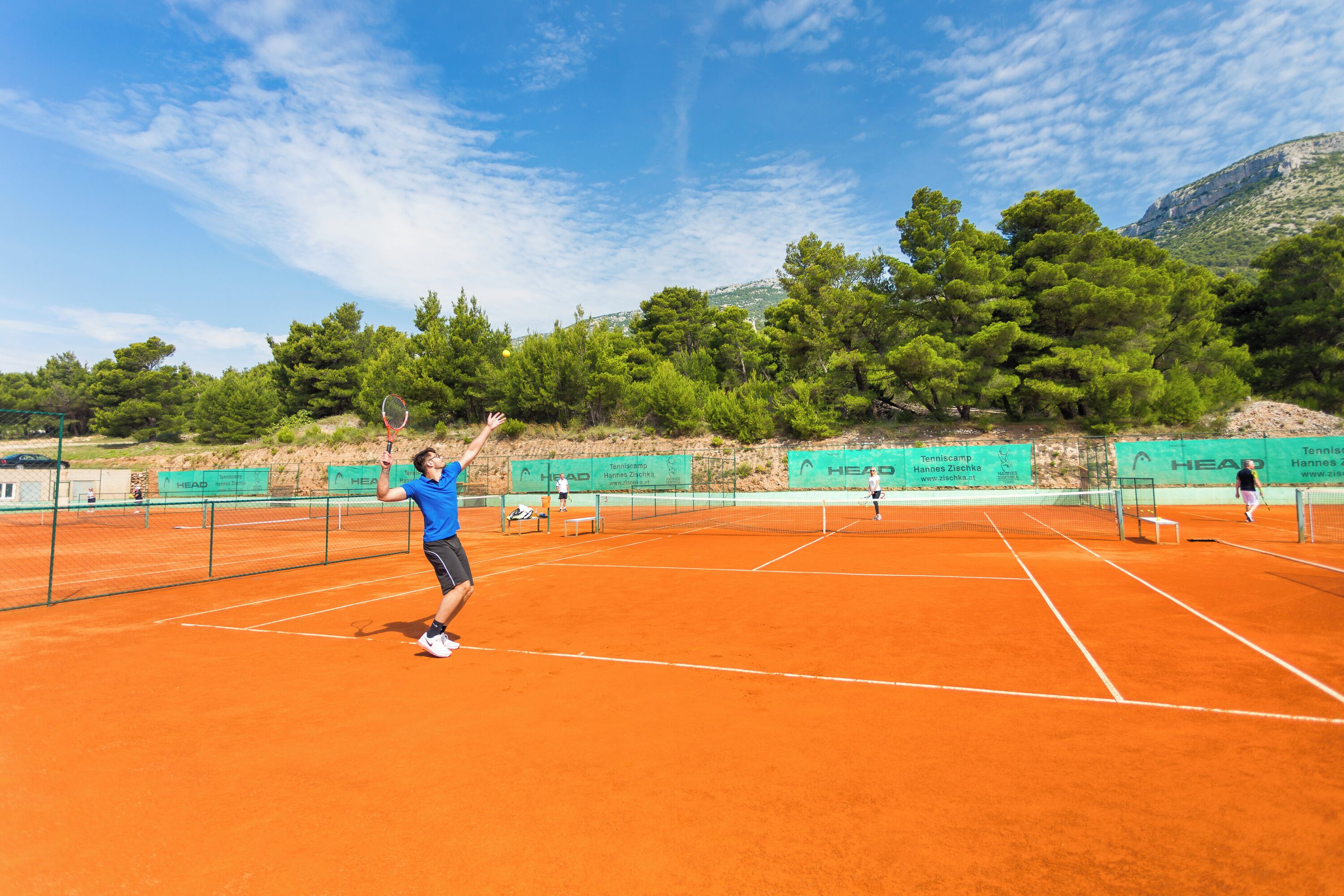 A tennis court with multiple courts, surrounded by trees and mountains.