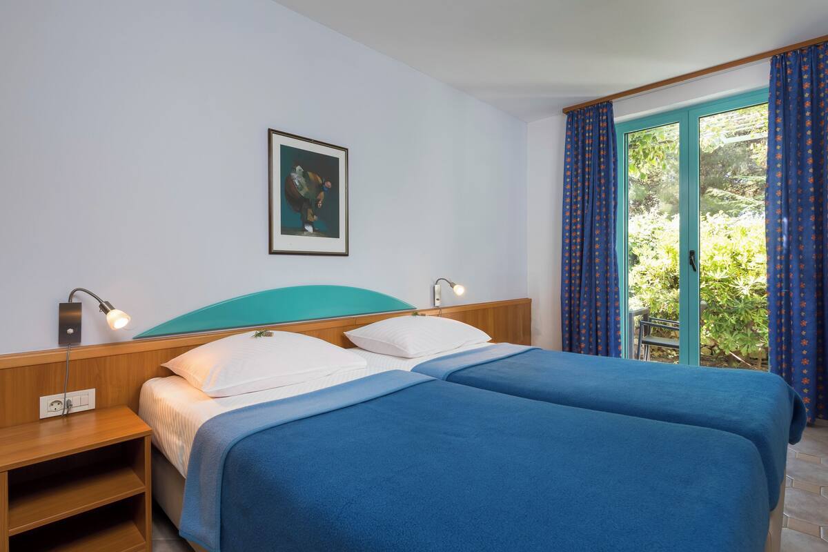 A double bed with a teal headboard, two bedside tables with lamps, a framed picture on the wall, and a view of greenery through a glass door.