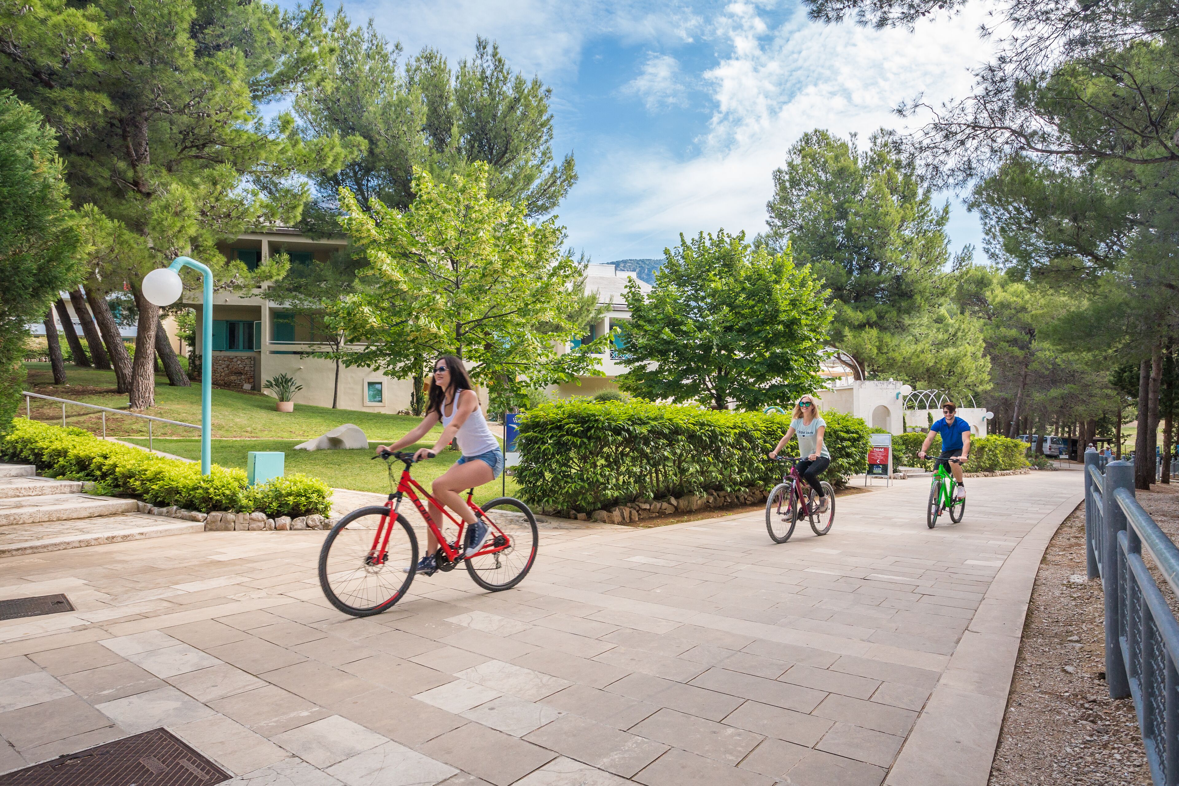 A paved pathway with three cyclists, surrounded by greenery and a building in the background.