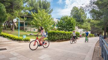 A paved pathway with three cyclists, surrounded by greenery and a building in the background.