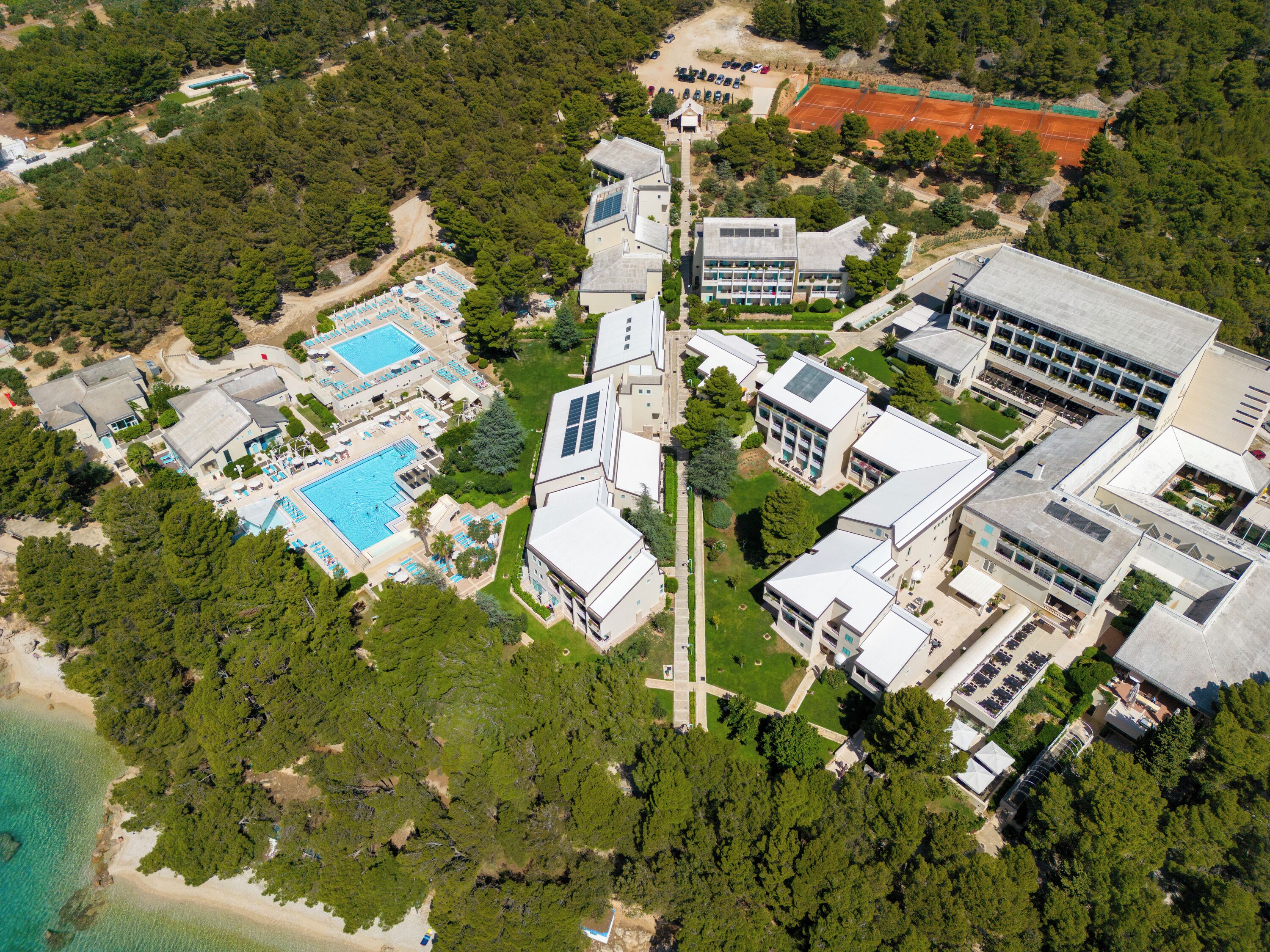 An aerial view of a resort with multiple swimming pools, a tennis court, and a central building with a cross-shaped driveway.
