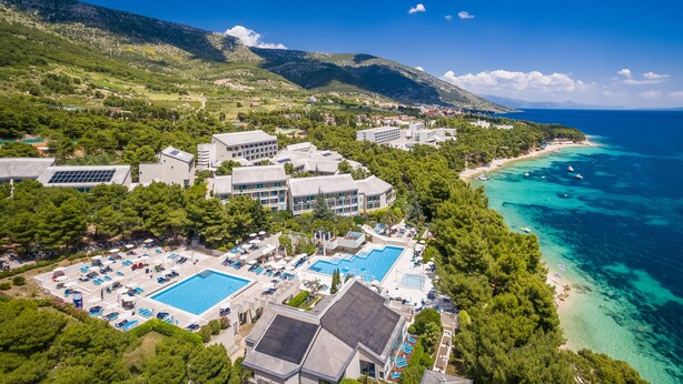 Vista aerea di un resort costiero con diversi edifici, una grande piscina e una spiaggia con acque cristalline color turchese.
