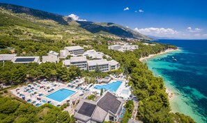 An aerial view of a coastal resort with multiple buildings, a large swimming pool, and a beach with clear turquoise waters. - Bretanide Sport & Wellness Resort (Bol)