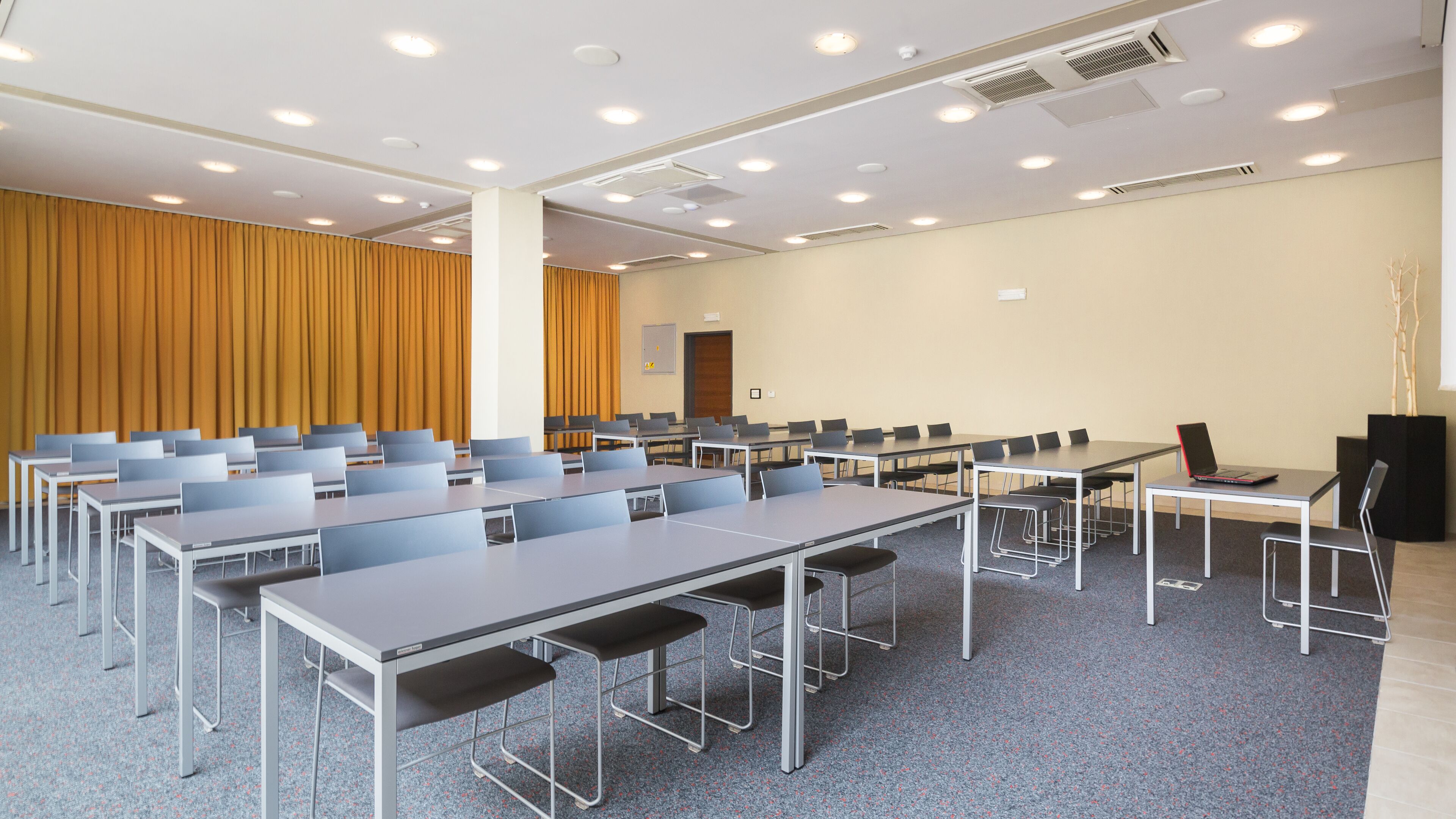 Un salón de conferencias con varias mesas y sillas, un telón grande y una puerta.