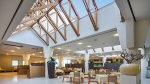 Un'ampia hall d'albergo con soffitto alto, luce naturale proveniente da lucernari, una reception centrale e diverse aree ristorazione.