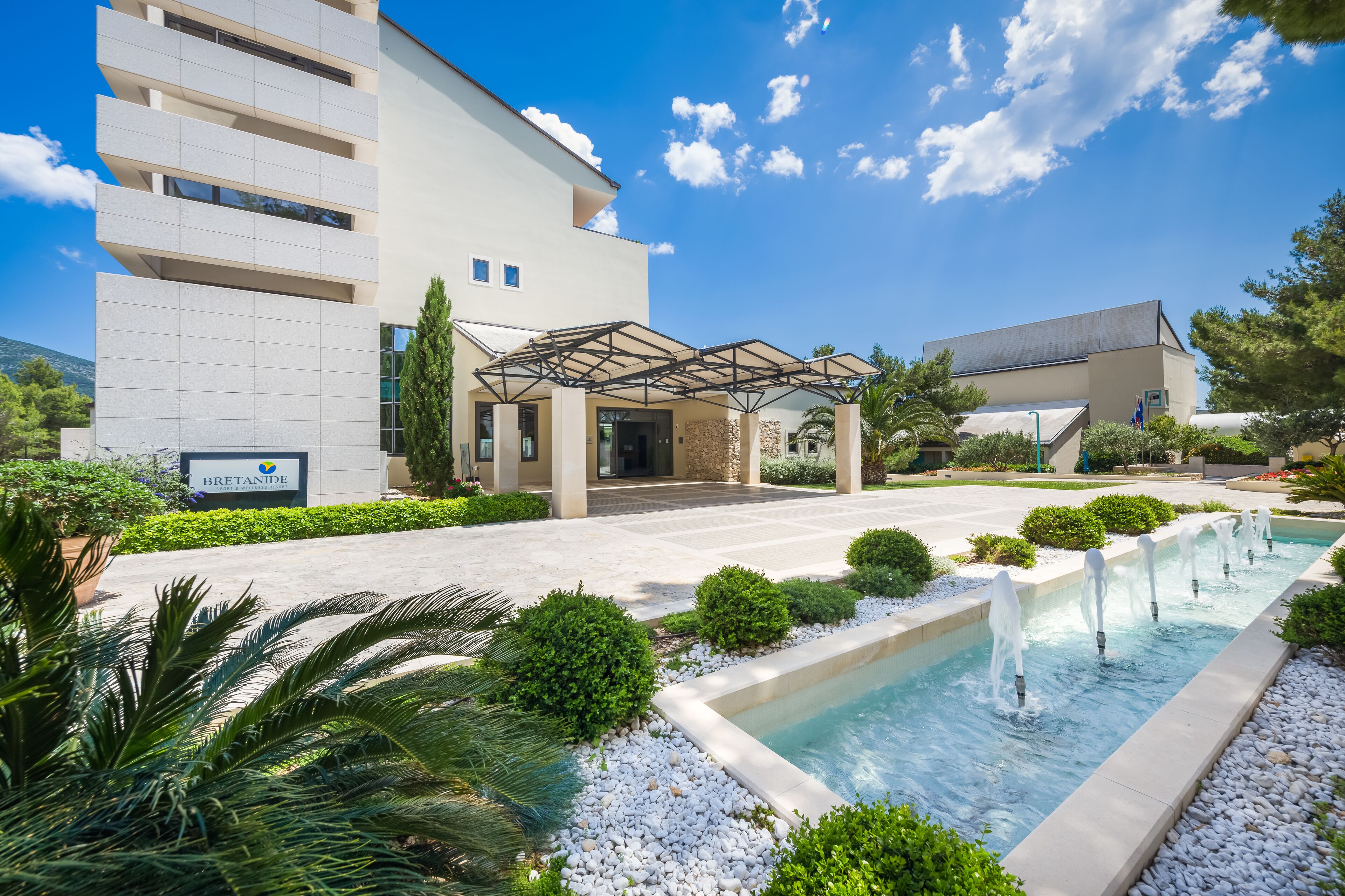 Un hotel moderno con entrada cubierta, piscina con fuentes y jardines bien cuidados.