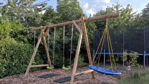 Un'altalena in legno con due sedie a dondolo e un trampolino blu in una zona boschiva.